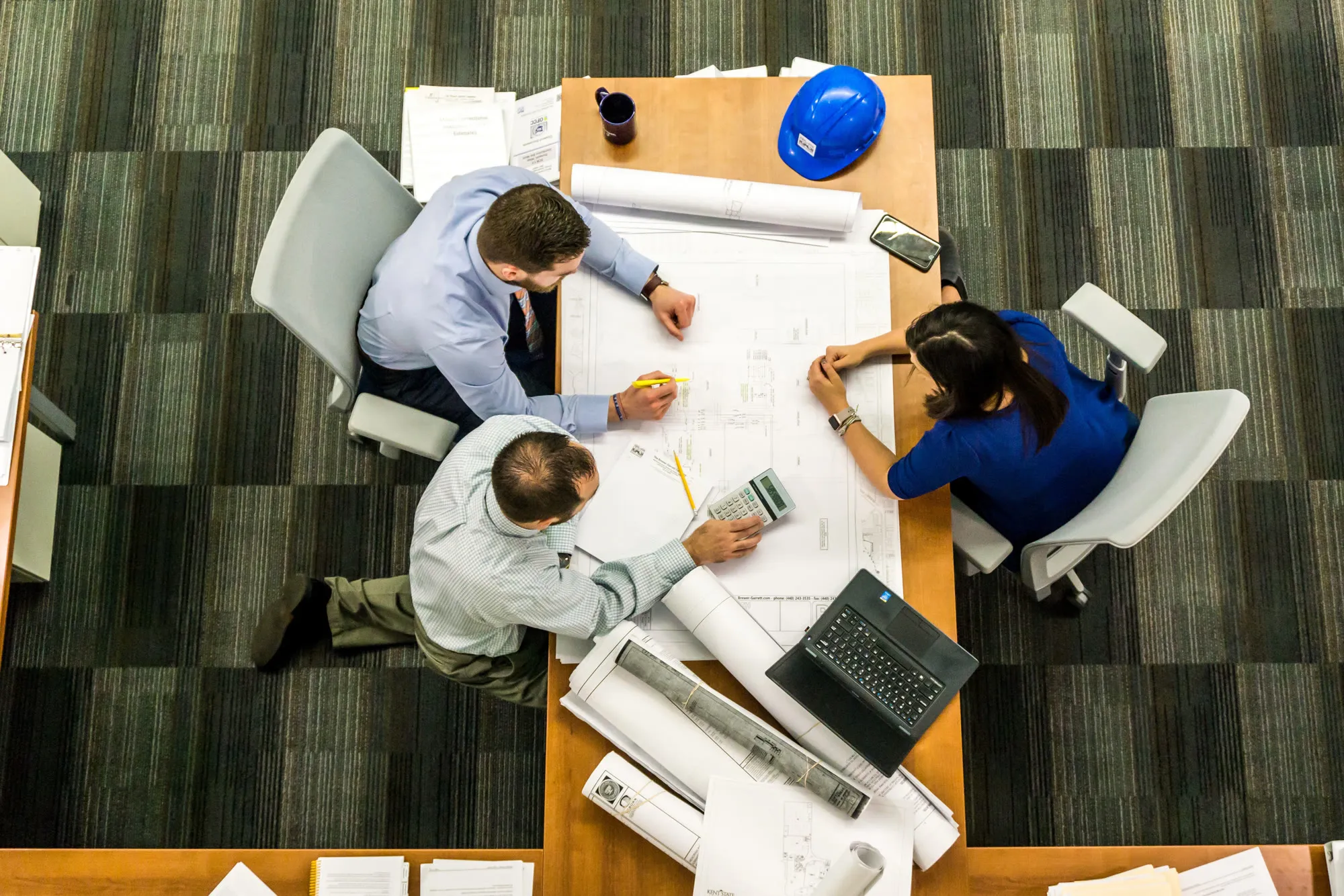 Tres profesionales revisando planos y documentación sobre una mesa de trabajo, con un casco de seguridad al lado