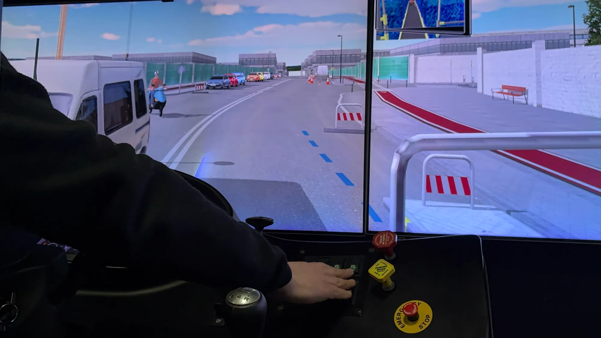 Vista desde la cabina del simulador: un conductor sujeta el volante frente a un escenario urbano con tránsito y barreras de obra
